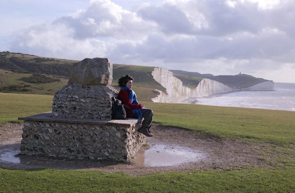 A woman sits on a stone bench with her eyes closed, facing the sea with the white cliffs of Seven Sisters clearly visible in the landscape behind.